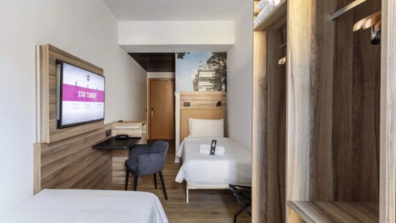 Hotel room at B&B Hotel Como Baradello with two single beds, wooden furniture, a mounted TV, chair, and open closet with hangers