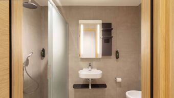 Modern bathroom at B&B Hotel Como Baradello with a shower, frosted glass partition, rectangular mirror, white sink, and soap dispensers