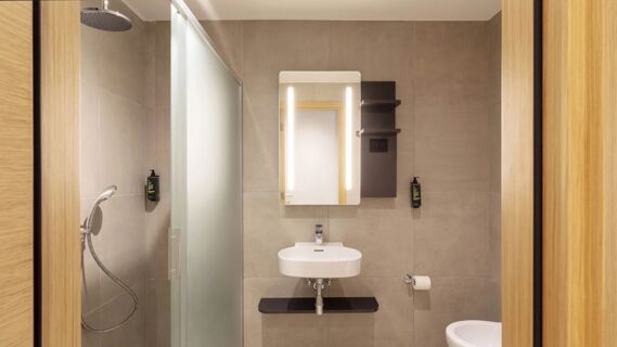 Modern bathroom at B&B Hotel Como Baradello with a shower, frosted glass partition, rectangular mirror, white sink, and soap dispensers
