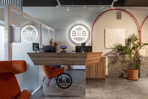 Reception desk with two Lenovo computers and orange chairs in a modern lobby of B&B Hotel Como Baradello with plants and tiled wall paneling