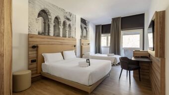 Hotel room at B&B Hotel Como Baradello with two beds, wooden accents, a TV, desk, chair, and large windows with curtains