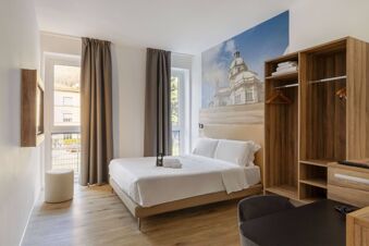 Hotel room at B&B Hotel Como Baradello with a large bed, wooden open wardrobe, two windows with beige curtains, and a wall mural of a cathedral