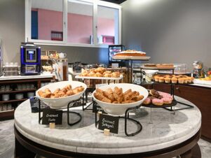 Breakfast buffet at B&B Hotel Quarto D'Altino with croissants, muffins, donuts, cakes, coffee machine, and fruit displayed on marble tables