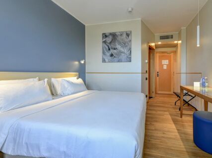 Modern hotel room with two beds, white linens, a blue wall, abstract wall art, wooden door, and table at B&B Hotel Quarto D'Altino