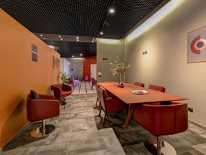 Interior view of meeting and lounge area with red chairs, a long orange table, plants, and abstract wall art at B&B Hotel Quarto D'Altino