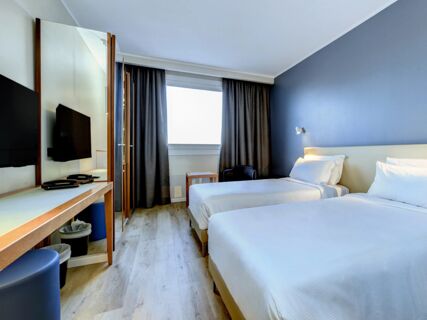 Two twin beds with white linens and pillows next to a blue wall and window in a room at B&B Hotel Quarto D'Altino