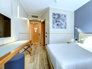 Modern hotel room at B&B Hotel Quarto D'Altino featuring a bed with white linens, wooden door, abstract wall art, and flat screen TV on the wall