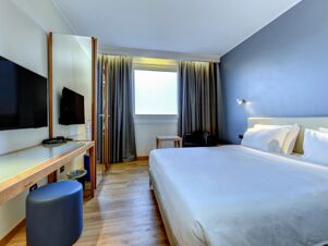 B&B Hotel Quarto D'Altino room featuring a bed with white linens, wall-mounted TV, full-length mirror, desk, chair, and window with curtains
