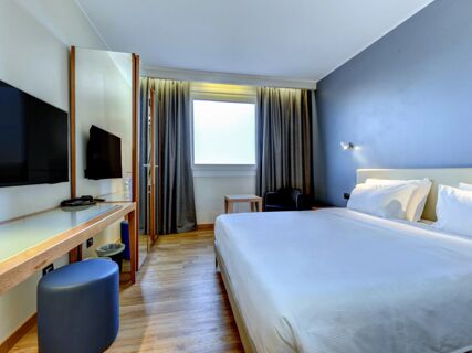 B&B Hotel Quarto D'Altino room featuring a bed with white linens, wall-mounted TV, full-length mirror, desk, chair, and window with curtains