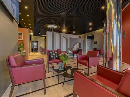 Interior lounge area of B&B Hotel Quarto D'Altino with red armchairs, black patterned cushions, small tables, and a dark grid ceiling