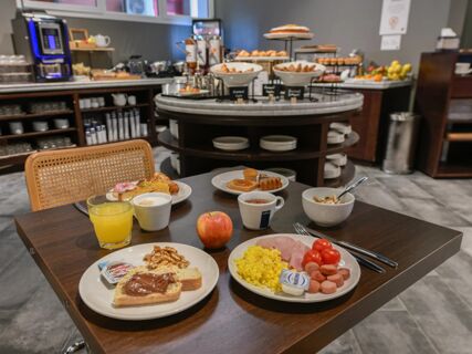 A breakfast spread with plates of scrambled eggs, ham, sausages, tomatoes, toast with Nutella, walnuts, orange juice, coffee, and an apple in the dining area of B&B Hotel Quarto D'Altino