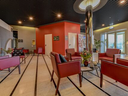 Lounge area with red leather chairs, small tables with flowers, a patterned floor, and glass doors at B&B Hotel Quarto D'Altino