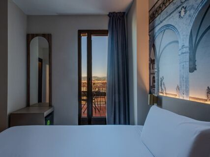 Modern hotel room at B&B Hotel Firenze Centro featuring a made bed, wooden framed mirror, balcony door, and blue curtains