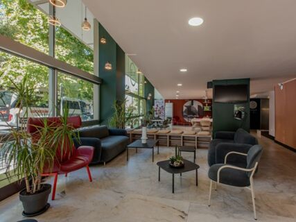 Seating area with red and grey armchairs, black sofas, potted plants, and tables in B&B Hotel Firenze Centro lobby with large windows