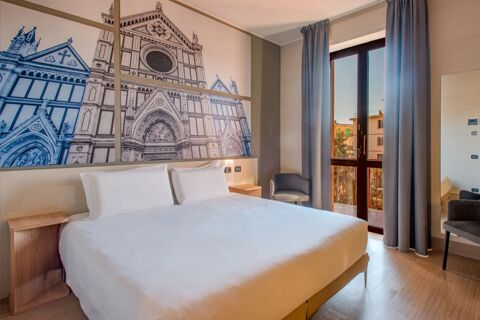 Hotel room at B&B Hotel Firenze Nuovo Palazzo di Giustizia with a bed, two pillows, bedside tables, chair, large window, and wall art of a cathedral facade
