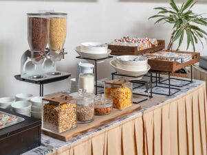 Breakfast buffet with cereal dispensers, bowls, milk, assorted packaged snacks, and a green plant at B&B Hotel Calenzano First