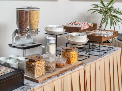 Breakfast buffet with cereal dispensers, bowls, milk, assorted packaged snacks, and a green plant at B&B Hotel Calenzano First