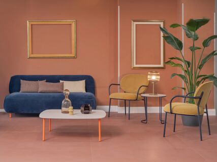 Interior of B&B Hotel Calenzano First with a blue sofa, beige and pink pillows, mustard chairs, empty gold frames, and green plant