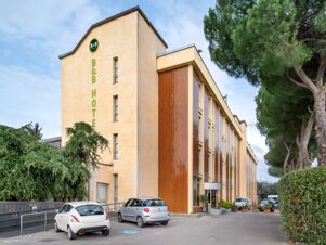 Exterior view of a beige and brown multi-story building with parked cars and greenery featuring B&B Hotel Calenzano First signage