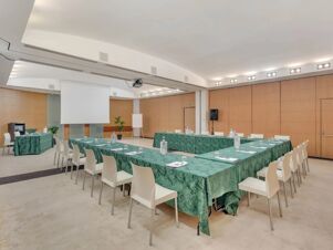 Conference room with green tablecloths on tables arranged in U-shape, white chairs, projector screen, and plants at B&B Hotel Calenzano First