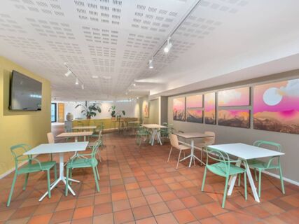 Dining area with white tables, green and beige chairs, terracotta floor tiles, and colorful wall art at B&B Hotel Alba
