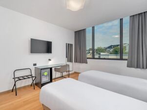 Hotel room at B&B Hotel Alba with two single beds, wooden floor, large window with curtains, desk, chair, wall-mounted TV, and luggage rack