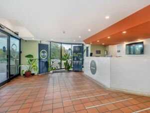 Reception area of B&B Hotel Alba with a white front desk, large windows, orange ceiling panel, and potted plants near entrance