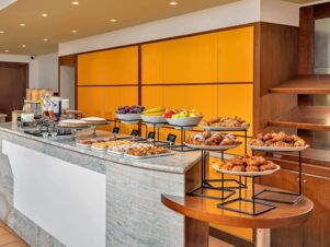 Buffet counter at B&B Hotel Alba with various pastries, bowls of fruit, cereals, and condiments against a yellow wall