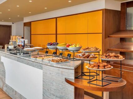 Buffet counter at B&B Hotel Alba with various pastries, bowls of fruit, cereals, and condiments against a yellow wall