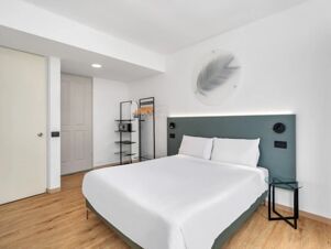Minimalist hotel room at B&B Hotel Alba with a double bed, white linens, green headboard, wooden floor, and shelves with hangers