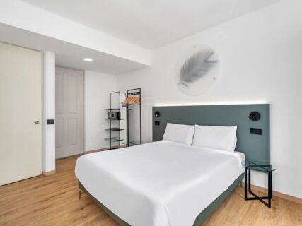 Minimalist hotel room at B&B Hotel Alba with a double bed, white linens, green headboard, wooden floor, and shelves with hangers