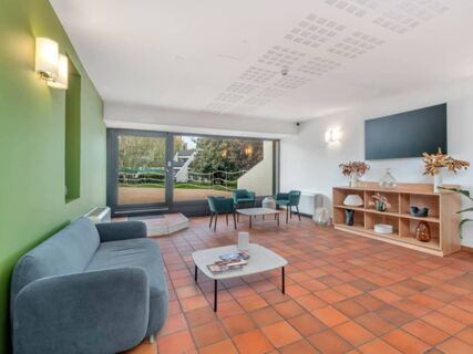 Modern lounge area at B&B Hotel Alba with blue armchairs, a sofa, a coffee table with magazines, and large windows showing greenery outside