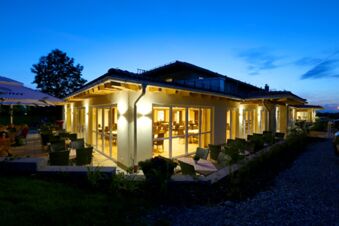 Au&szlig;enaufnahme des beleuchteten Hotel-Restaurant-Cafe Friedenseiche bei Abendd&auml;mmerung mit Terrasse und Sitzgelegenheiten im Freien.