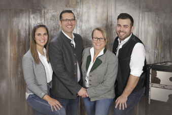 Vier l&auml;chelnde Personen in Trachtenjacken vor einer rustikalen Holzwand, Bild f&uuml;r das Team des Hotel-Restaurant-Cafe Friedenseiche.