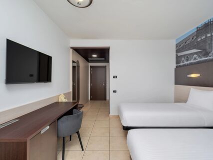 Room interior at B&B HotelPark Hotel Mondov&igrave; with two white beds, a wall-mounted TV, wooden desk, and chair on beige tile flooring