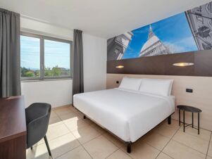 Minimalist hotel room at B&B HotelPark Hotel Mondov&igrave; with a large bed, two bedside lights, a desk, chair, and window with curtains
