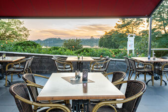 Outdoor seating area with wooden tables and wicker chairs under a red canopy at Hotel am Gothensee by a lake view at sunset