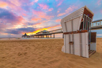 A wicker beach chair on sandy shore with a pier and buildings at sunset sky visible near Hotel am Gothensee