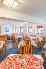 Dining area inside Hotel am Gothensee with wooden chairs, tables covered with red and white tablecloths, and windows with red frames