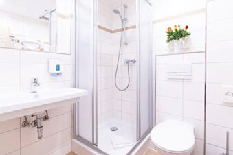 Bathroom with white tiled walls, walk-in shower, sink, toilet, and flower decoration at Hotel am Gothensee