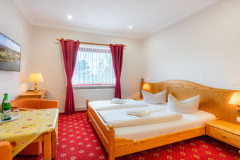Hotel am Gothensee room featuring a double wooden bed with white linens, red curtains, red carpet, table with a bottle, and wall-mounted TV