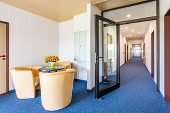 A seating area with four beige armchairs around a small table holding a vase of sunflowers in Hotel am Gothensee hallway with blue carpet