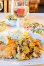 Fried fish with a lemon wedge, roasted potatoes, and salad served on a white plate with a glass of white wine at Hotel am Gothensee