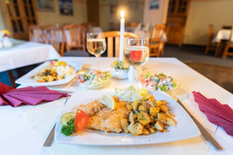 Fried fish with roasted potatoes, lemon wedge, and vegetables on a table at Hotel am Gothensee with wine glasses and candle