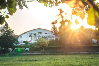 Hotel am Gothensee building surrounded by green trees and grass field under bright sunlight with sun rays visible