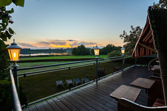 View of a sunset over a lake with green fields and lounge chairs from a wooden terrace at Hotel am Gothensee with lamps on