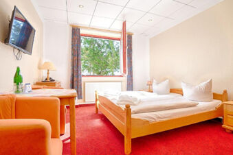 Hotel am Gothensee room with red carpet, wooden bed and furniture, orange chairs, a table, and a window showing green trees