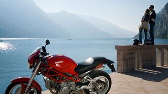 赤いドゥカティ・モーターバイクが山の前の湖岸に佇み、背景にはカップルがいる。