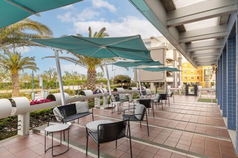 Outdoor seating area with black metal chairs, small tables, large blue umbrellas, palm trees, and buildings at B&B Hotel Chioggia Airone