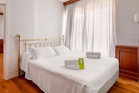 Double bed with white linens and pillows, folded towels on top, sheer curtains, and wooden flooring inside B&B Hotel Chioggia Airone